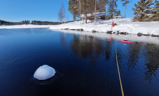 Vinterlandskap med ett rep över öppet vatten, där en person med röda kläder står på andra sidan vattnet.