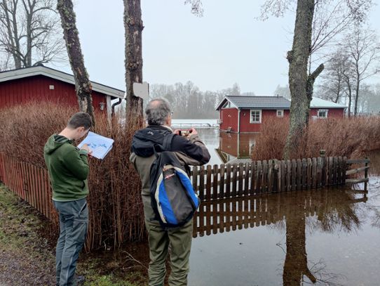 På bilden syns hur Roxen har översvämmat ett sommarstugeområde. Fredrik jämför den verkliga översvämningen med en översvämningskarta, medan Göran fotar översvämningen. 