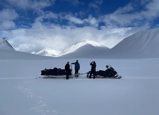 Konturerna av tre människor och två skotrar syns i fjällmiljö. Marken och de höga bergen är vita av snö. 