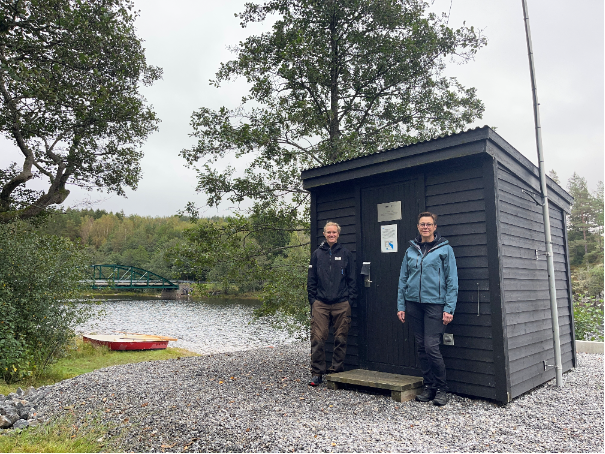 Två kvinnor utanför en mörk byggnad med vatten och en eka i bakgrunden. 