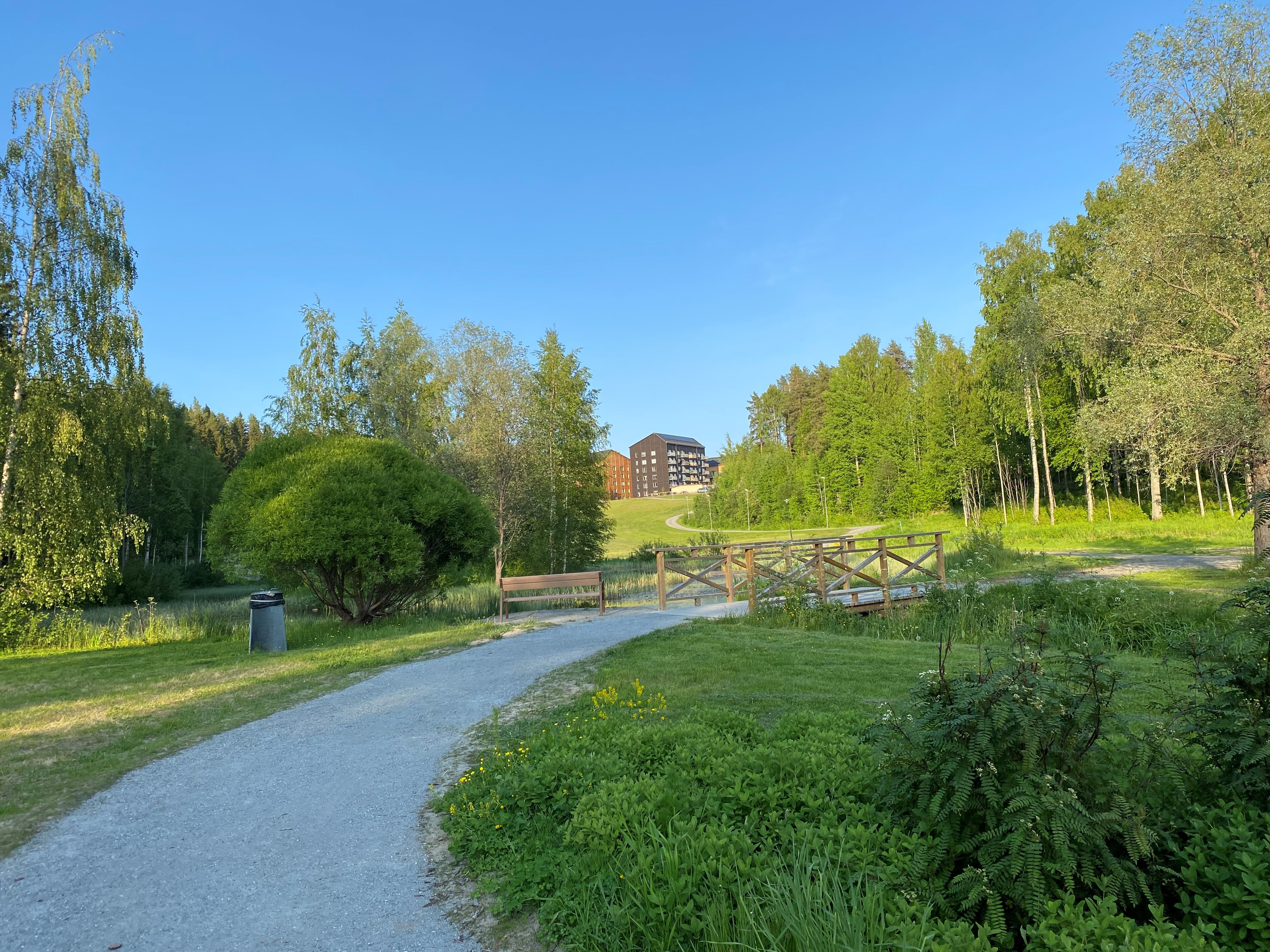 Grön
parkmiljö med gångväg, bro och rik vegetation, med bostadshus i bakgrunden.