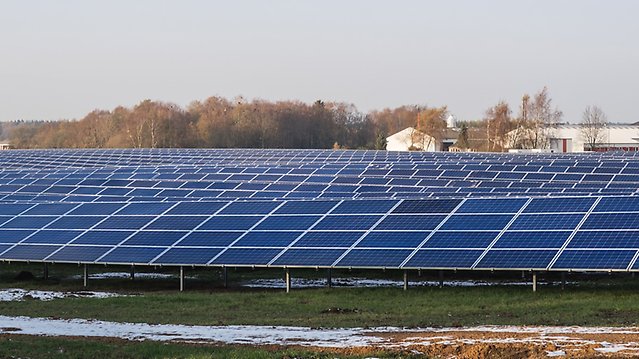 Solar panels in long rows on open land, with industrial buildings in the background.