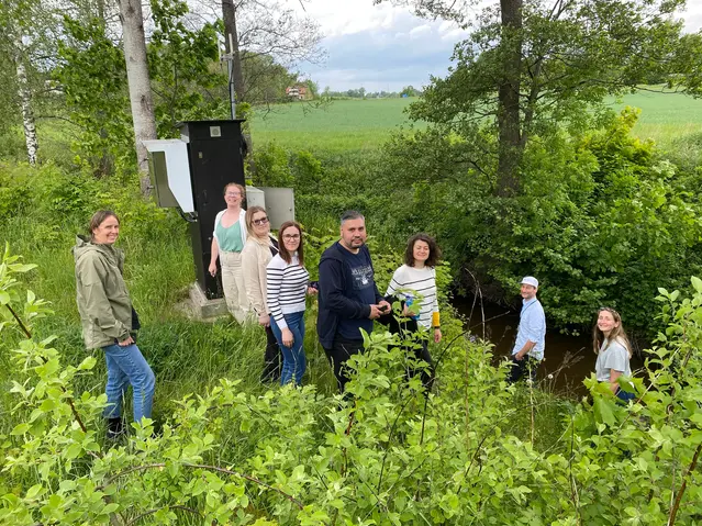 A group of eight people standing in the greenery by a stream, smiling at the camera.