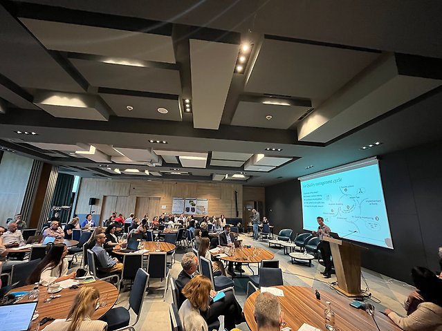 Konferensdeltagare sitter vid runda bord i en konferenssal och lyssnar på en presentation. På en stor skärm visas en bild om luftkvalitetsarbete.