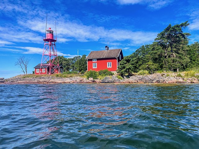 Rödmålad fyr och stuga i sommarsol, omgivet av blå vågor och blå himmel.