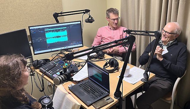 Tre personer runt ett bord med mikrofoner och datorer, de samtalar och ser glada ut. 