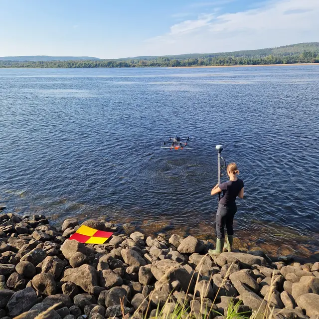 A girl is standing with her back towards the camera by a large river. A drone is flying to the left.