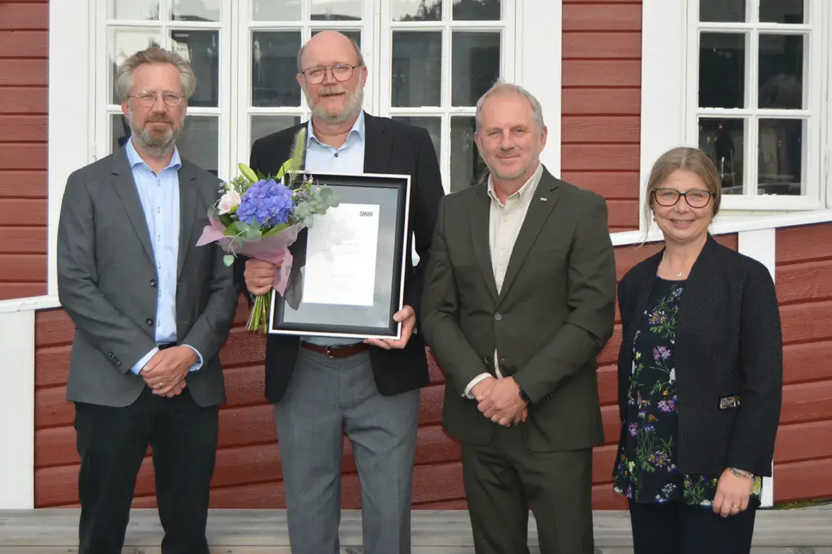 Fyra personer står uppradade utomhus varav den ena håller ett inramat diplom i handen.