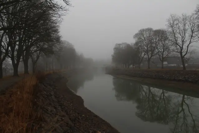 Göta Kanal under låga ljusgrå startusmoln, med sin karaktäristiska trädallé på var kanalbank. De nakna träden speglas i den blanka vattenytan. I mitten av bilden syns Malforsbron. Sikten är begränsad av dis.