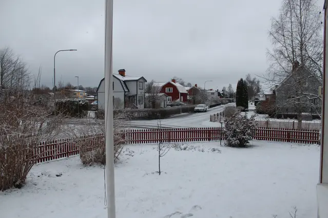 Snö över ett villkvarter under en gråvit stratustäckt himmel, ett trädgårdshörn i förgrunden omgivet av staket. Bortom staketet en gatukorsning och i bakgrunden ett antal villor
