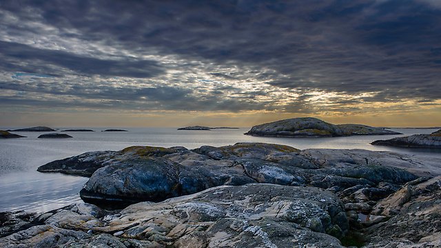 Klippor och små öar vid havet under en dramatisk kvällshimmel med mörka moln och ljus som bryter igenom i horisonten.