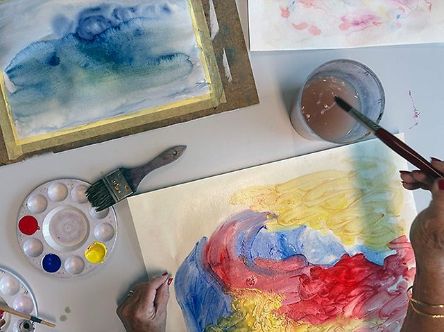 Bild på akvarellmålningar i blått, gult, rött och en hand som håller i en pensel som doppas i vatten. 