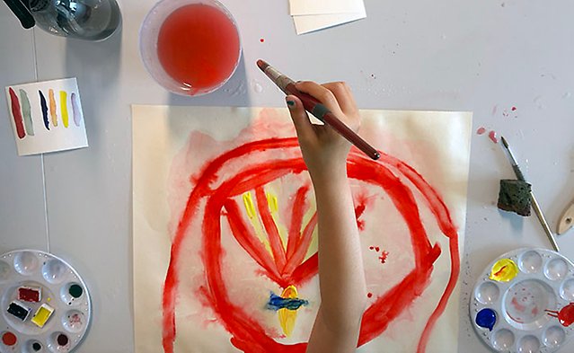 En liten hand håller i en pensel över en akvarellmålning som går i mest rött, men även i gult och blått. 