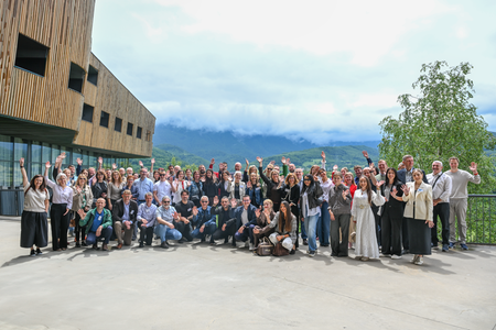 Gruppfoto från konferensen i Sarajevo. Ett stort antal deltagare står samlade utomhus framför en byggnad och vinkar mot kameran. I bakgrunden syns grönskande berg och molnigt väder.