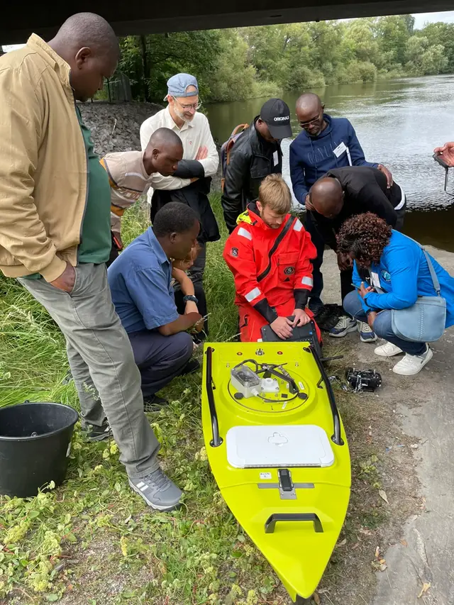 Nio personer står och tittar på ett hydrologisk mätinstrument. De är vid ett vattendrag.