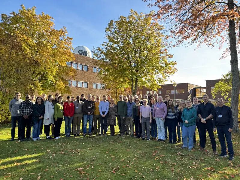 Gruppbild med ett 40-tal personer tagen utomhus. Personerna ler mot kameran. I bakgrunden syns träd med höstfärger och SMHIs kontor.