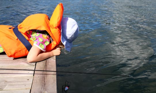 Ett barn iklädd flytväst ligger på en brygga och tittar ner i vattnet