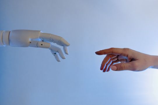 Robothand sträcker sig mot hand mot en ljusblå bakgrund.