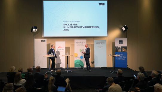 Bild på konferensens scen, där Markku Rummukainen och moderator Johan Kuylenstierna pratar med varandra inför publiken. 