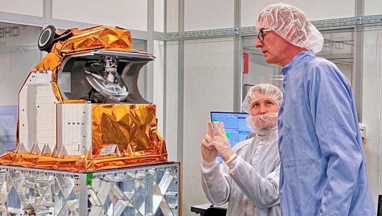 Två män iklädda skyddskläder tittar på en satellit i ett labb. 