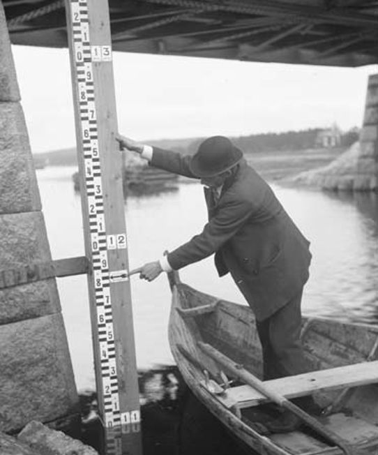 Gråvit bild på man i båt i vattendrag som pekar på en stor linjal på en stenbro.